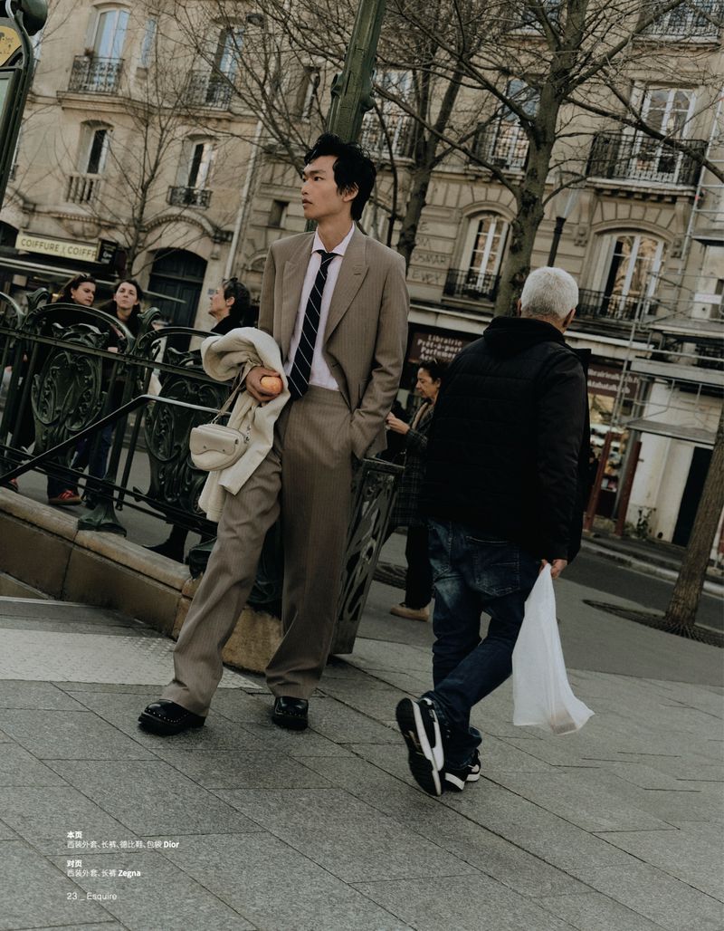 Man in a suit stands on a city street near a subway entrance. Buildings, pedestrians, and trees are visible in the background.