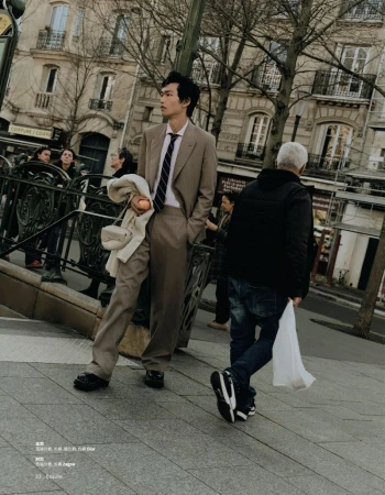 Man in a suit stands on a city street near a subway entrance. Buildings, pedestrians, and trees are visible in the background.