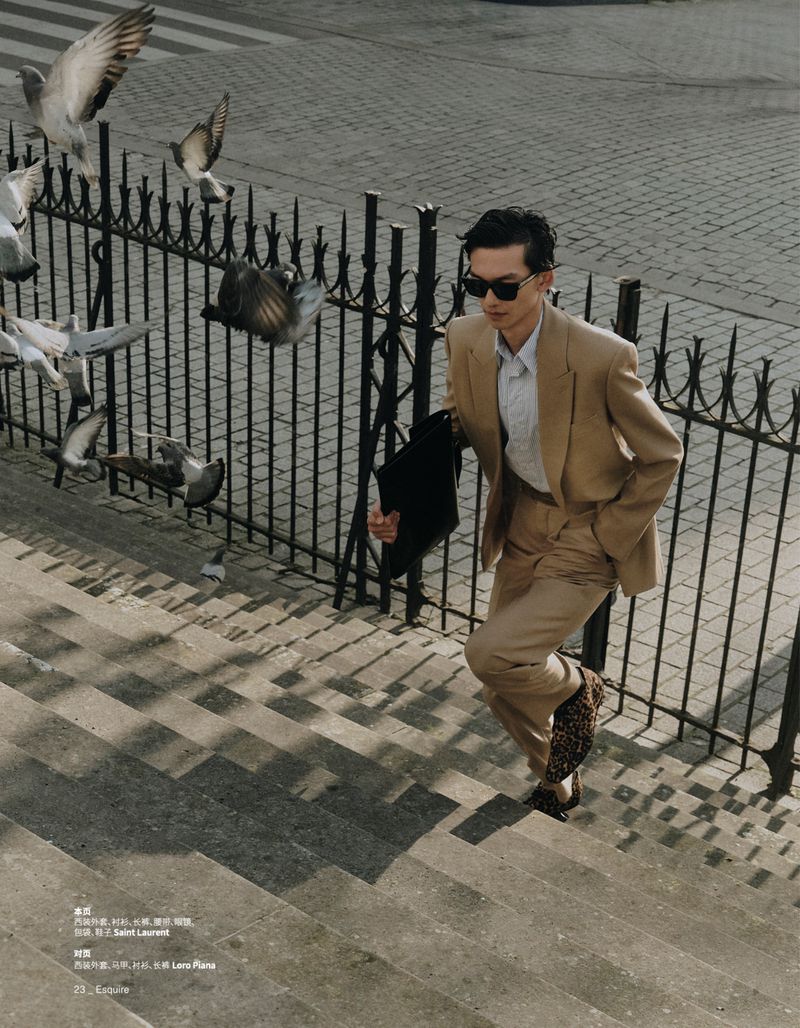 An adult male in a tan suit walks up gray stairs with iron railings, holding a black coat, surrounded by flying pigeons.
