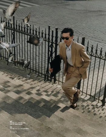 An adult male in a tan suit walks up gray stairs with iron railings, holding a black coat, surrounded by flying pigeons.