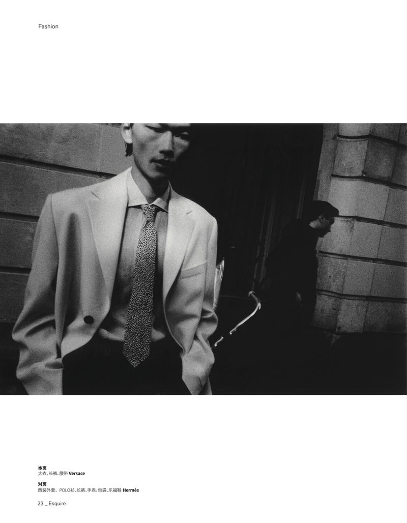 Black and white editorial fashion portrait of a man in a tailored blazer and tie standing on a city street.