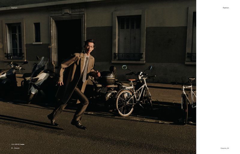 Fashion editorial shot of a man in a trench coat running on a street with scooters and bikes parked alongside a building in dim lighting.
