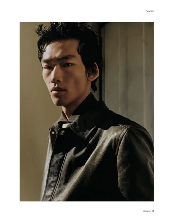 Portrait of a young adult Asian man in a brown leather jacket in a medium shot with soft, diffused lighting against a plain background.