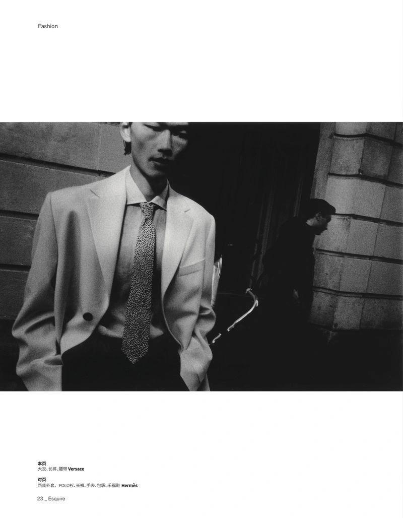 Black and white editorial fashion portrait of a man in a tailored blazer and tie standing on a city street.