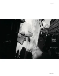 Monochrome fashion editorial image of a model wearing a tailored suit and tie, holding a small handbag on a city street.