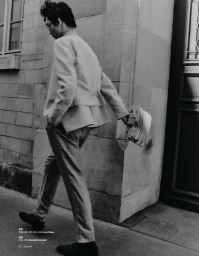 Monochrome full-length editorial shot of a man wearing a light suit jacket and trousers holding a newspaper on a stone street.