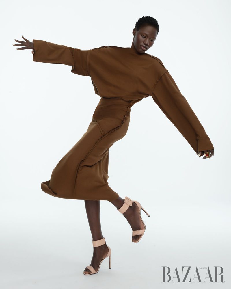 Fashion shot of an African-descent adult female posing in a brown outfit with beige high heels on a white backdrop in studio lighting.