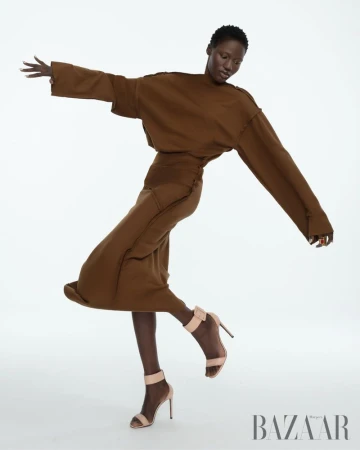 Fashion shot of an African-descent adult female posing in a brown outfit with beige high heels on a white backdrop in studio lighting.