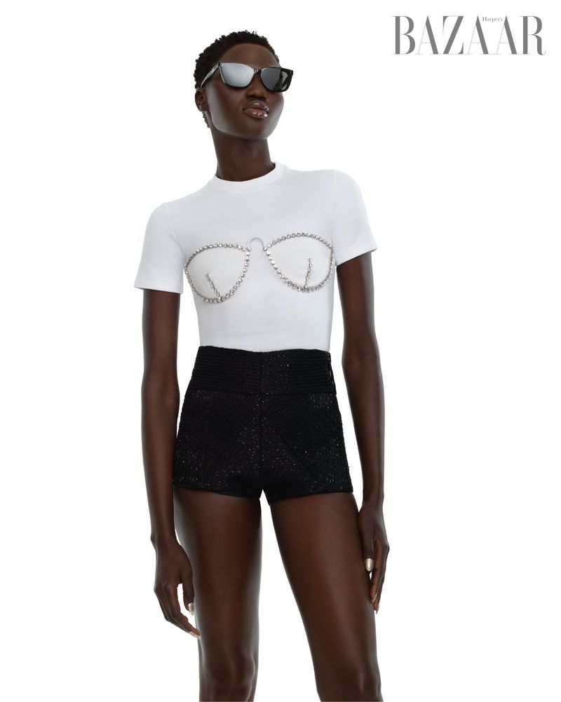 Fashion model wearing a white top with decorative bra design and black shorts poses in a studio with a white background.