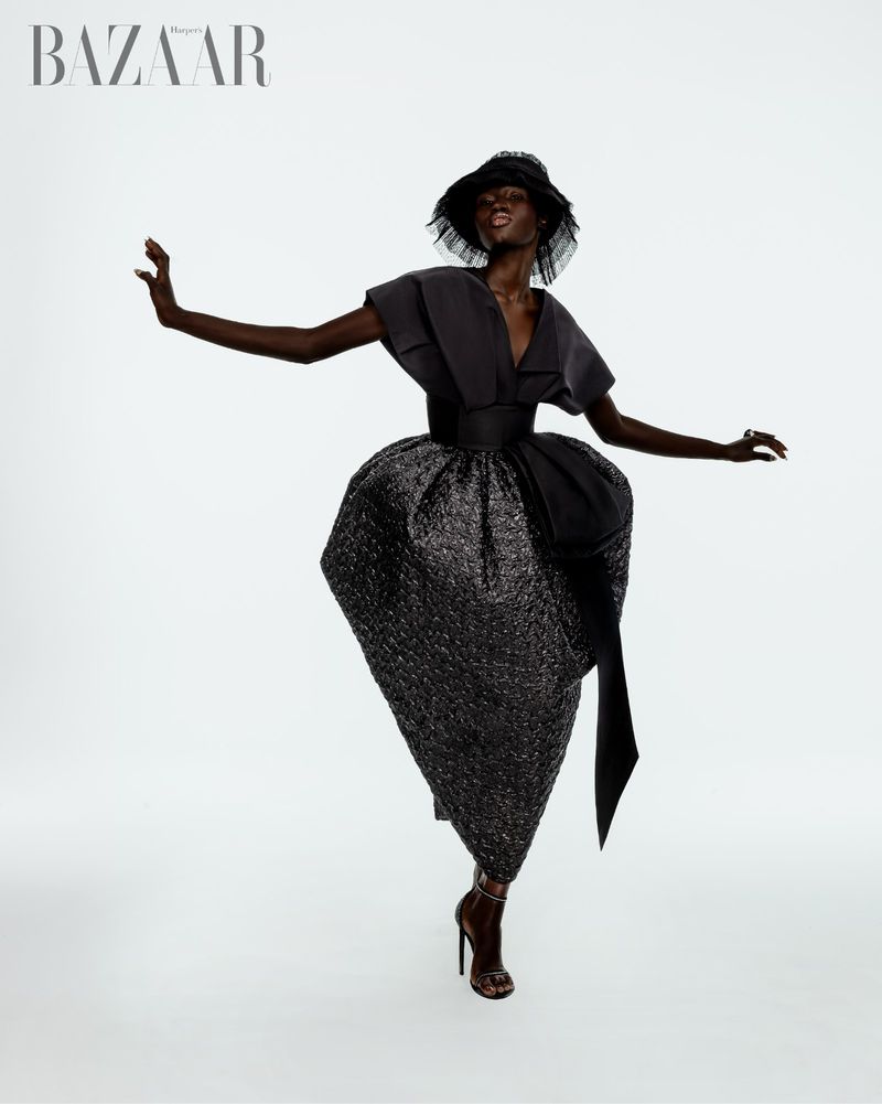 An elegant Black woman wearing a dramatic couture dress, hat, and high heels, striking a dynamic pose in a studio shot against a white background.