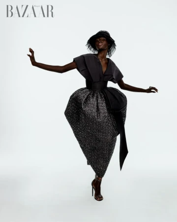 An elegant Black woman wearing a dramatic couture dress, hat, and high heels, striking a dynamic pose in a studio shot against a white background.