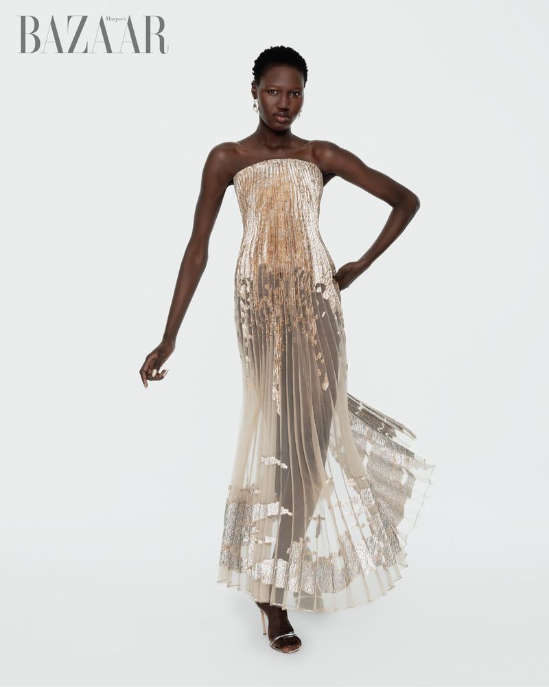 An elegant Black woman models a sleeveless, gold-and-beige embellished gown in a full shot in a bright studio setting for a fashion editorial.