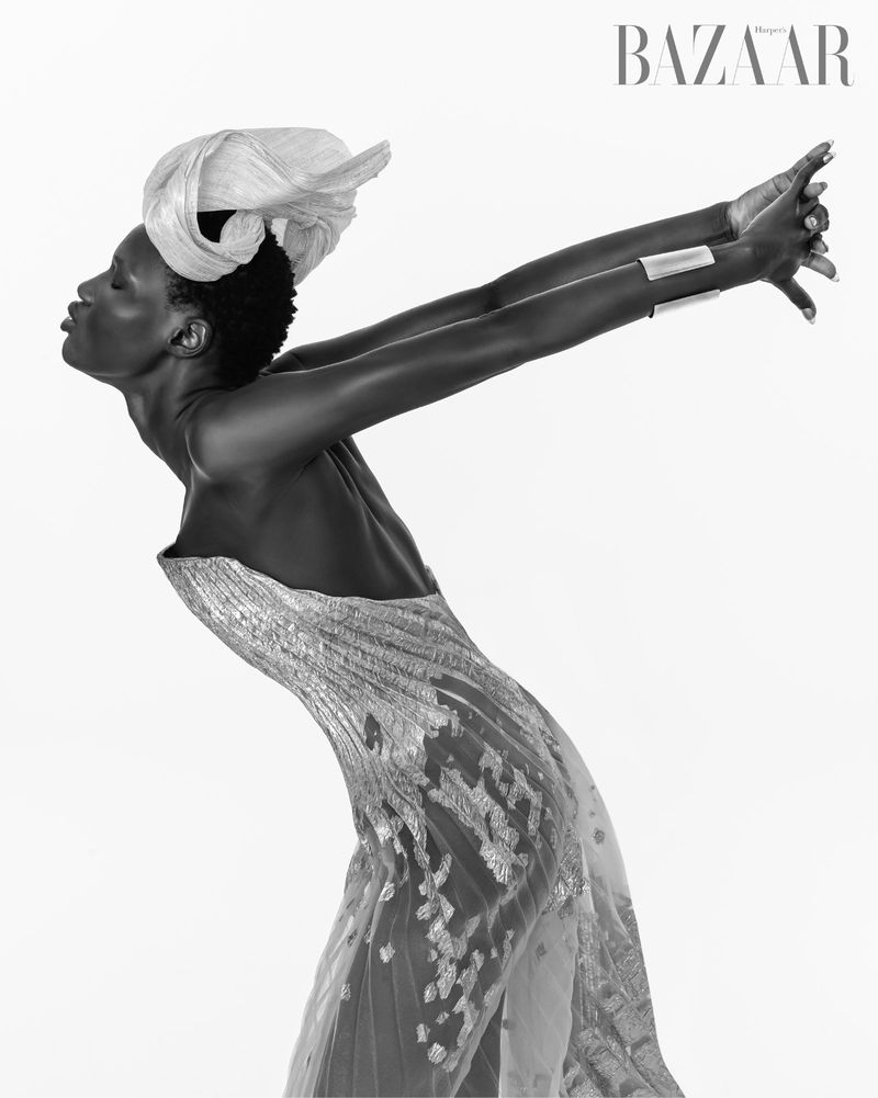 Elegant black and white fashion shot of a Black woman in a shimmering gown and headwrap posing in a white background. Bazaar logo visible.