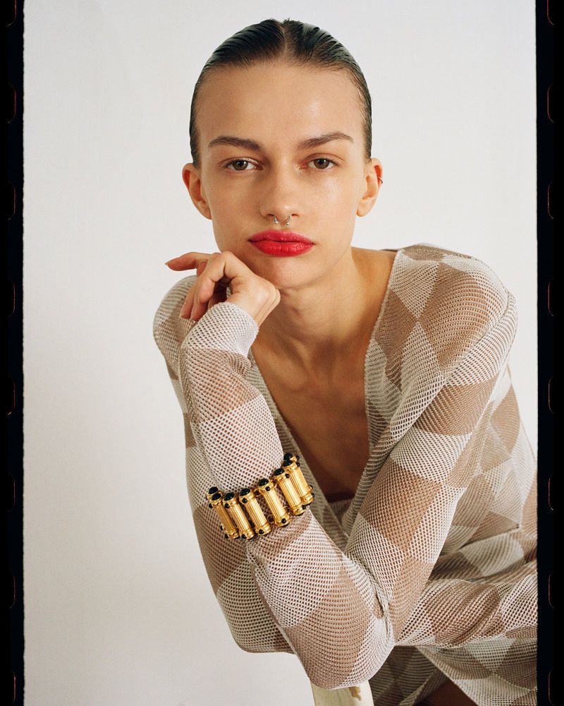 Headshot of a woman with red lipstick and slicked-back hair. She wears a mesh top, gold bracelet, and a septum piercing, in a studio.