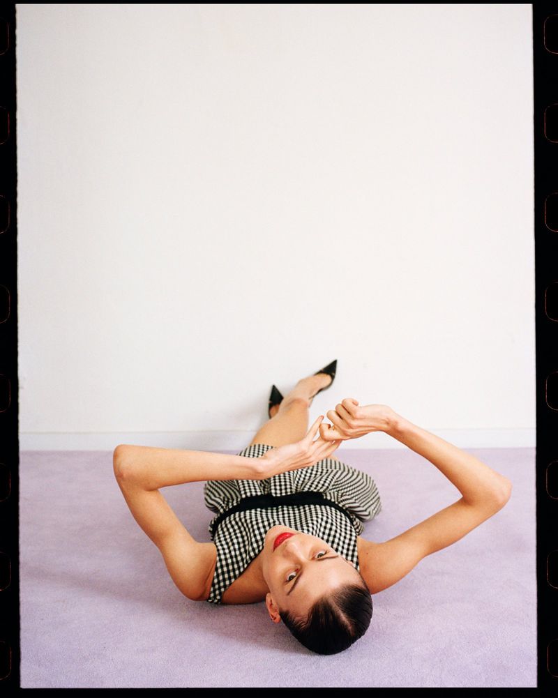 Woman in a gingham dress and black heels lies on a lilac carpet in front of a white wall, shot from an eye-level angle, indoor setting.