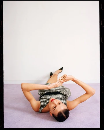 Woman in a gingham dress and black heels lies on a lilac carpet in front of a white wall, shot from an eye-level angle, indoor setting.