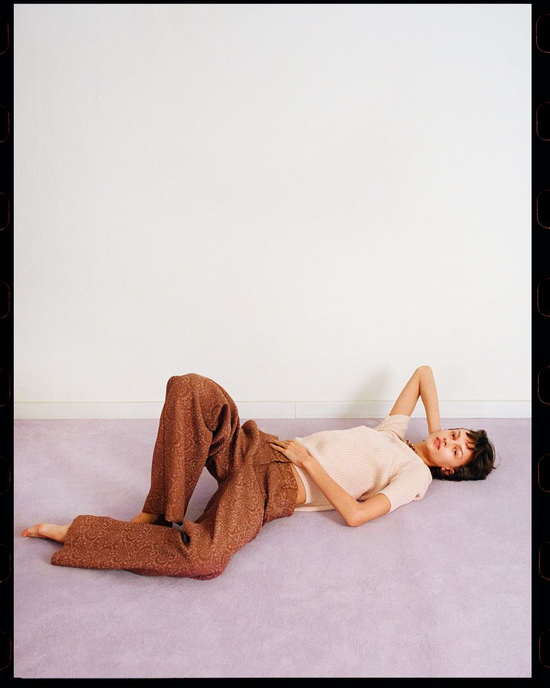 A young woman lies on a lavender carpet wearing a beige top and patterned brown pants in a minimal studio setting with white walls.