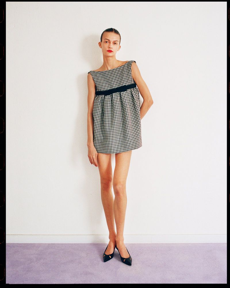 A woman models a black and white gingham mini dress with a black sash against a simple white wall with a lavender baseboard.