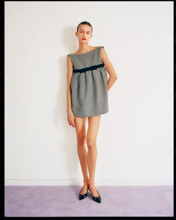 A woman models a black and white gingham mini dress with a black sash against a simple white wall with a lavender baseboard.