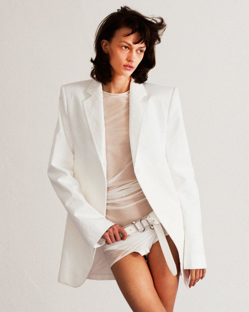 Brunette woman poses in studio wearing a white blazer and white shorts with a sheer top and white belt against a white background.