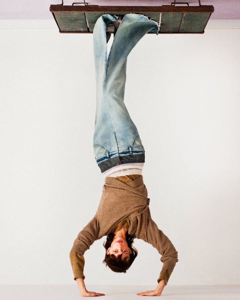 Inverted fashion editorial image featuring a model doing a handstand wearing light denim jeans and a brown sweater against a white wall.