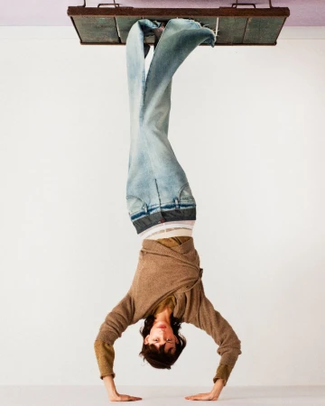 Inverted fashion editorial image featuring a model doing a handstand wearing light denim jeans and a brown sweater against a white wall.