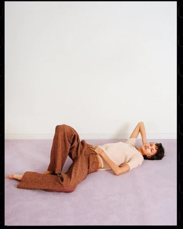 Model lying on a purple carpet wearing brown patterned trousers and a light top against a white wall.