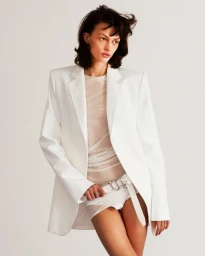 Model wearing a white blazer, sheer top, and white shorts against a neutral background for A Part Publications.