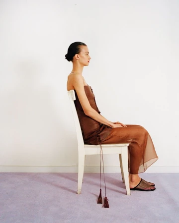 Model seated sideways on a white chair wearing a sheer brown dress against a plain wall