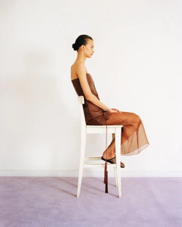 Model seated in profile on a white wooden chair wearing a brown dress against a plain wall and lavender floor