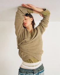 Woman in an olive green knitted cardigan and trousers with hands raised against the ceiling on a white background