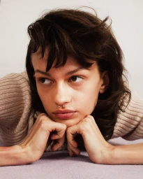 Close-up portrait of a woman resting her chin on her hands with dark hair and septum piercing against a neutral background.