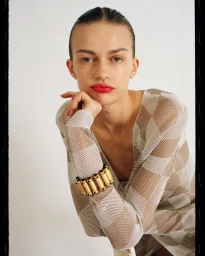 Model with slicked-back hair wearing a patterned top and chunky gold bracelet with red lipstick against a white background