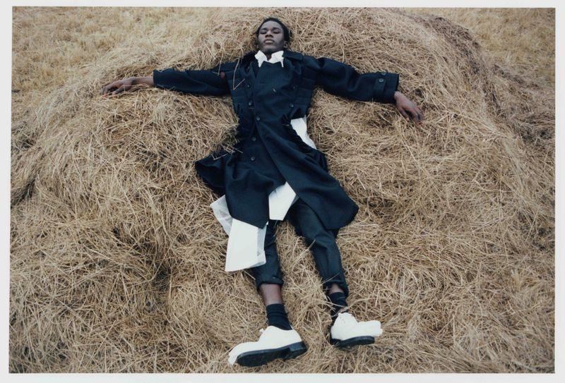 A person lies on a haystack, wearing a black coat, white shirt, dark pants, and chunky white shoes, outdoors in soft, diffused light.