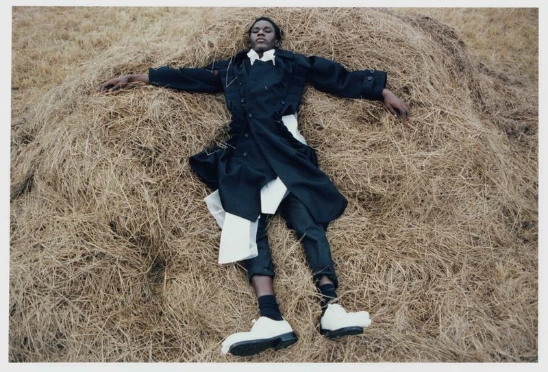A person lies on a haystack, wearing a black coat, white shirt, dark pants, and chunky white shoes, outdoors in soft, diffused light.