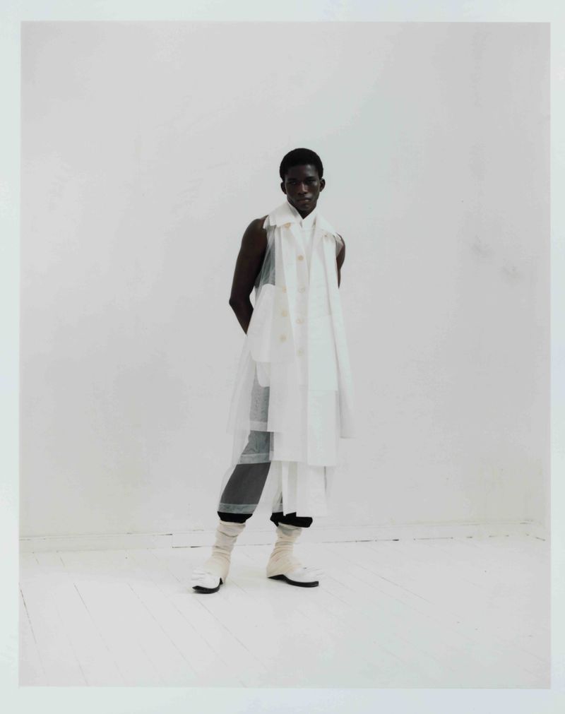 Full-length portrait of a Black male fashion model in a white layered outfit against a white backdrop in a studio setting, stylish and posed.