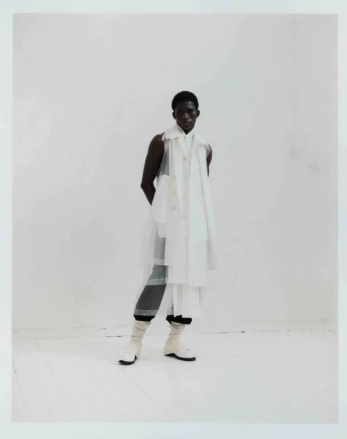 Full-length portrait of a Black male fashion model in a white layered outfit against a white backdrop in a studio setting, stylish and posed.