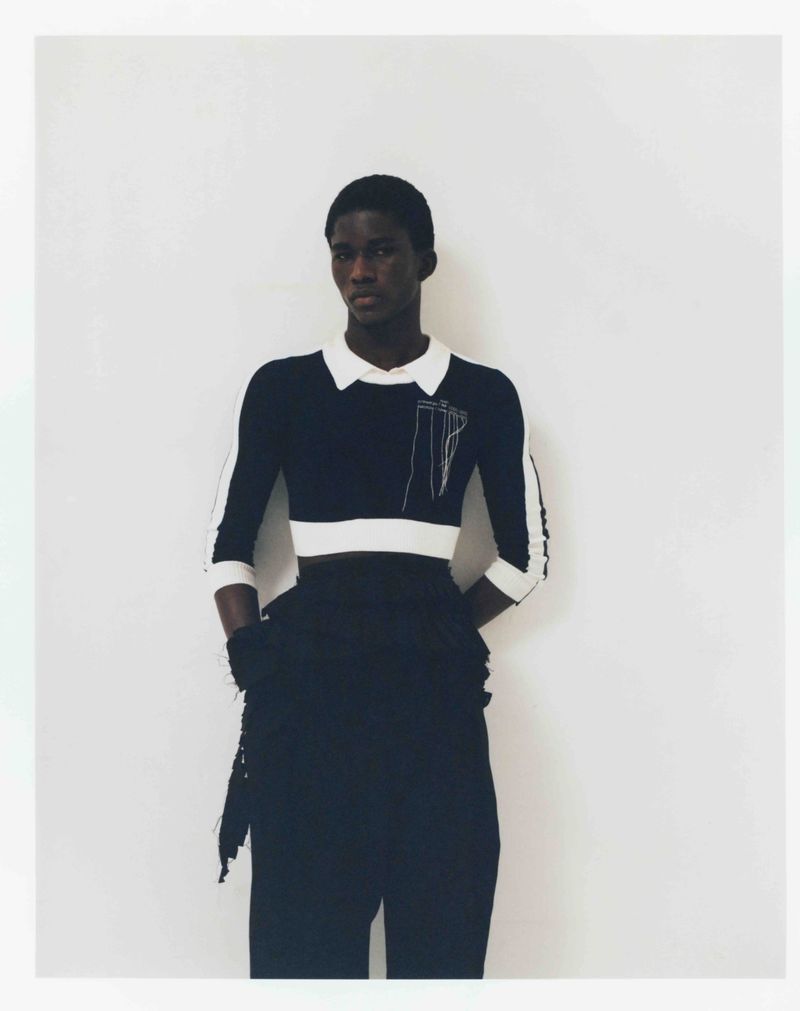 Portrait of an African American adult male in a black and white long sleeve shirt and black pants, posing against a white wall.