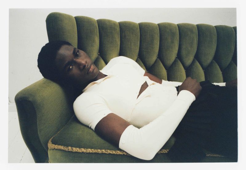 An adult male lies on a green velvet couch wearing a cream-colored shirt and black pants, looking at the camera in an indoor setting.