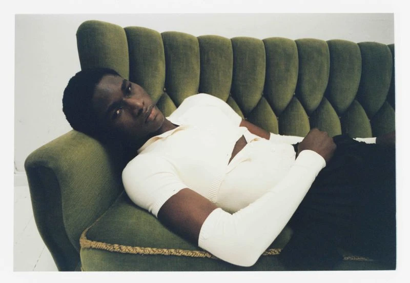 An adult male lies on a green velvet couch wearing a cream-colored shirt and black pants, looking at the camera in an indoor setting.