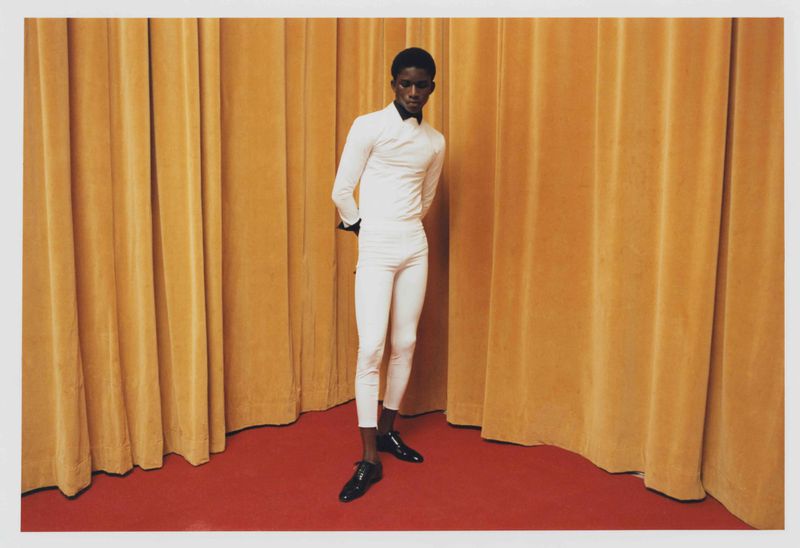 A Black male fashion model stands in a white outfit against a gold curtain with red carpet in a studio setting, striking a pose.