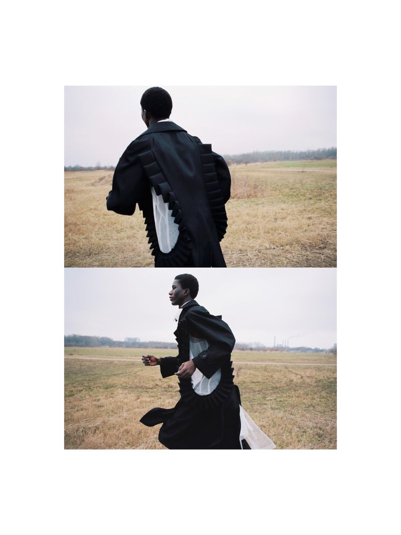 Young adult in black coat with flowing white accent runs in open field under overcast sky in fashion editorial shot. Low angle.