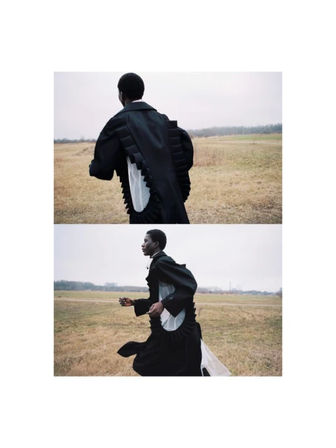Young adult in black coat with flowing white accent runs in open field under overcast sky in fashion editorial shot. Low angle.
