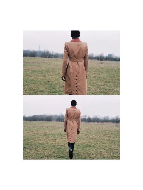 An African-descent adult female stands in a field, wearing a trench coat. The woman faces away from the camera in a rural environment.