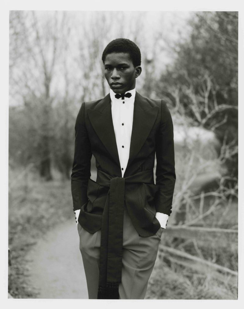 Young man in a black tuxedo with hands in pockets, standing on a path with blurred trees in the background, formal black-and-white portrait.