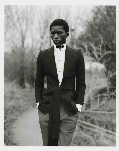 Young man in a black tuxedo with hands in pockets, standing on a path with blurred trees in the background, formal black-and-white portrait.