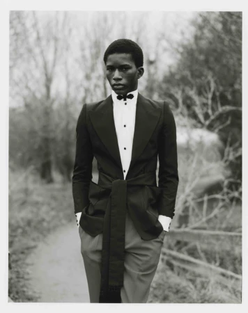 Young man in a black tuxedo with hands in pockets, standing on a path with blurred trees in the background, formal black-and-white portrait.