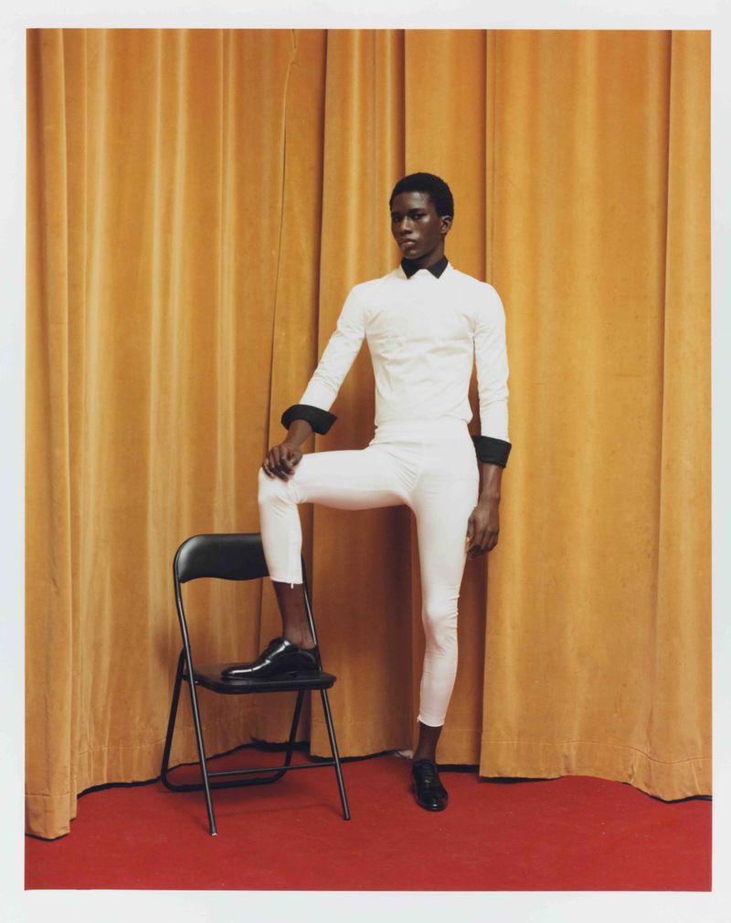 Young black male model in a white outfit poses with a black chair against a yellow curtain and red floor in a studio.
