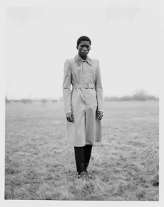 Model Mohammed Banze wearing a long belted trench coat standing in a field for The Greatest Magazine editorial by Paw Gissel.