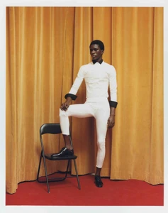 Model wearing white shirt and trousers standing with one foot on a folding chair before yellow curtains.